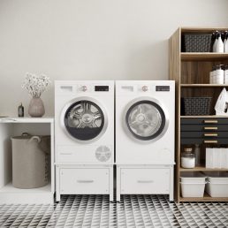 Een moderne wasruimte met een witte wasmachine en droger naast elkaar op een wasmachine verhoger met lade, opbergplanken met manden en flessen, een wasmand, een vaas met bloemen en geometrische zwart-witte vloertegels.