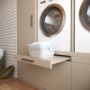 Een rieten wasmand met opgevouwen witte handdoeken staat op een uitschuifbare plank onder een voorlader wasmachine in een moderne wasruimte met beige kasten.